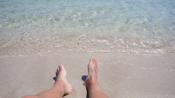 Close-up of a Man's Legs Lying on a Sandy Beach in Azure Sea Water, Small Waves Beating Against His