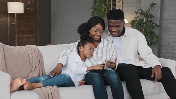 African Family Talk Laugh Looking at Smartphone Using Funny Apps Sit on Couch Choosing Online