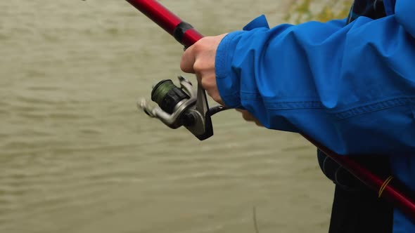 Close-up of a fisherman in a blue jacket spinning a reel handle on a red rod