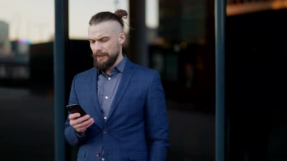 a Mustachioed and Bearded Man in a Plaid Jacket Stands with a Phone in Hand and Looks at the Screen