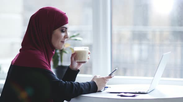 Attractive Muslim Girl with Hijab on Her Head is Drinking Cappuccino While Searching for Something