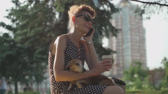 A Mature Pet Owner of a Dachshund Dog Lounges on a Bench Drinks Coffee to Herself and Talks on the