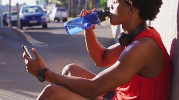 Fit african american man exercising in city taking a break, using smartphone, sitting drinking water
