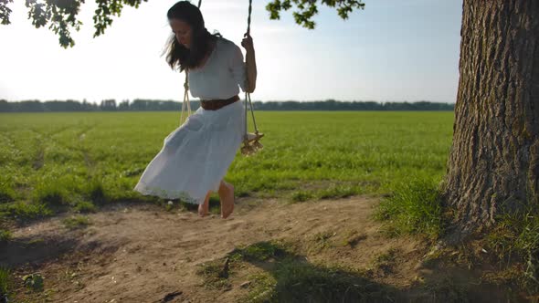 Young Barefoot Girl in a Beautiful White Rustic Dress Is Swinging on a Swing in the Open Field.