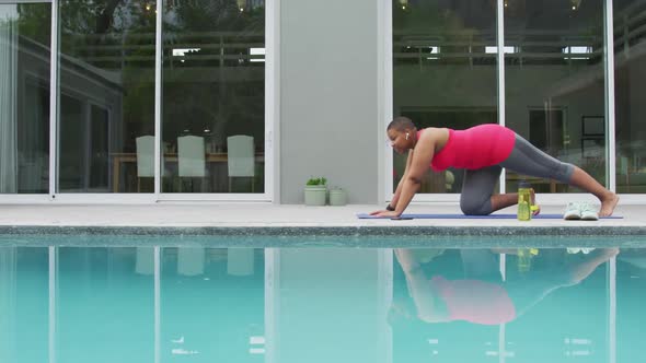 Happy african american plus size woman practicing yoga, stretching next to swimming pool in garden