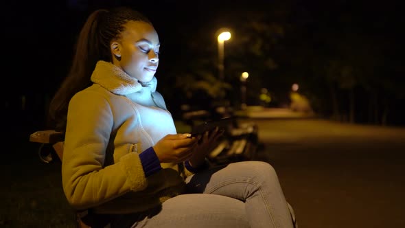 A Young Black Woman Looks at a Tablet with a Smile As She Sits on a Bench in a City Park at Night