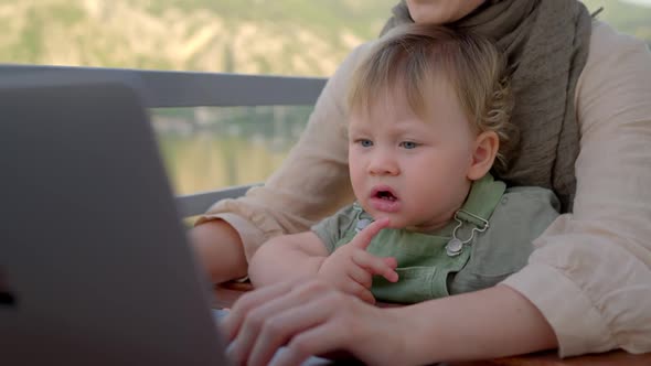 Mother Multitasking Holding Baby Infant and Using Computer Laptop at Home