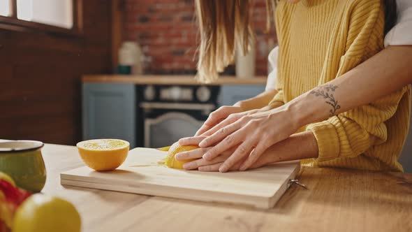 Unknown Mother Helping Her Child to Cut Orange on Wooden Board with Knife Standing By Table in