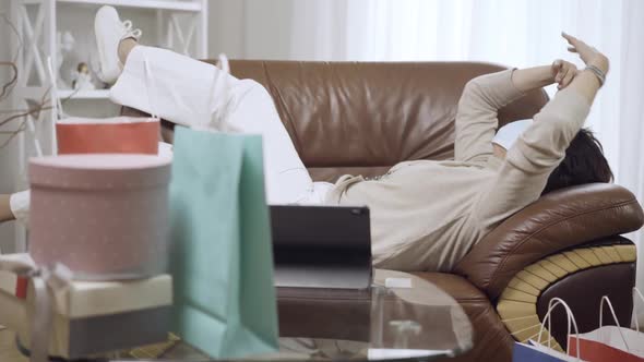 Carefree Caucasian Middleaged Woman Lying on Couch in Covid Face Mask with Shopping Bags and Tablet