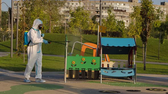A Man in a Protective Suit and a Respiratory Gas Mask Disinfects the Slides on the Playground