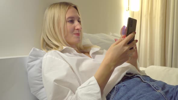 a woman at home reads a text message on her smartphone while lying on the bed.