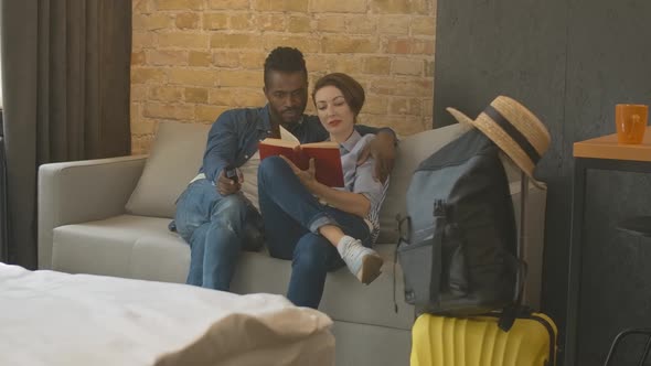 Wide Shot of Relaxed Interracial Couple Sitting on Couch with Remote Control and Book. Packed