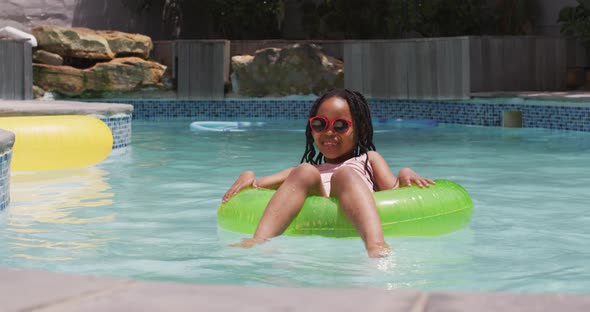 Happy african american girl wearing sunglasses floating on inflatable ring in swimming pool