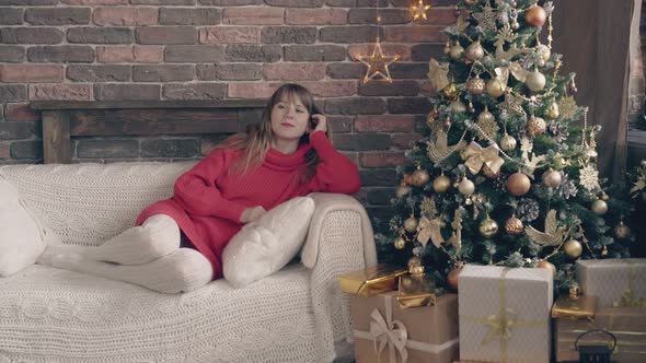 Woman in Red Dress Sweater Lies on Sofa Near Christmas Tree