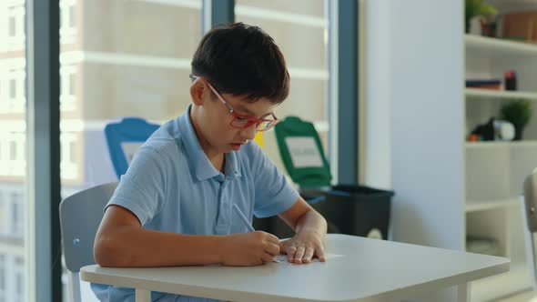 Asian Pupil Wearing Glasses Sits at a Desk at School and Fills Out Tests