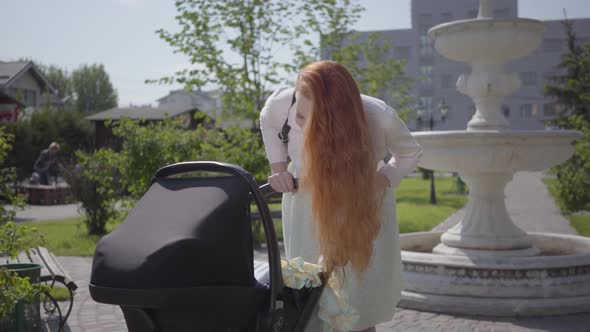 Adorable Red-haired Mom Bent Over a Pram and Smiling in the Park