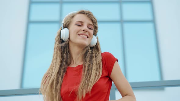 A Young Girl in Headphones is Listening to Music and Amusingly Dancing on a City Background