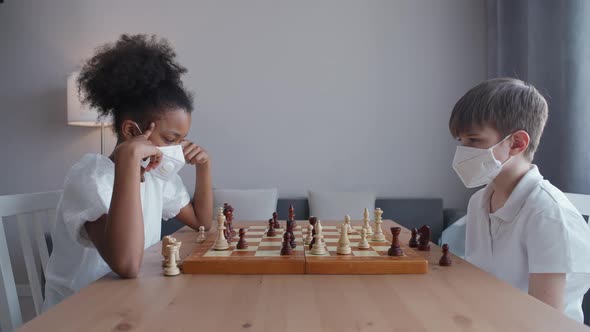 Children in Medical Mask on Selfisolation an African Teen Girl and a Caucasian Boy Play Chess