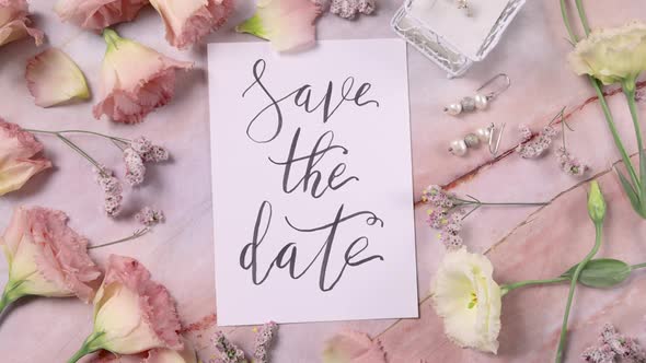 SAVE THE DATE card on a marble table near pink flowers top view zoom in