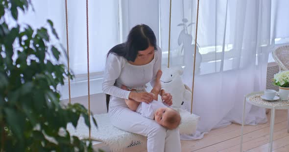 Pretty and Caring Mother Rocking Her Newborn Baby on a Swing at Home a Bright Room