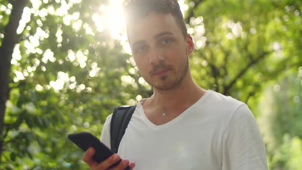 Handsome Young Man Stands in the Park Inside a Tunnel of Trees with a Phone in His Hands