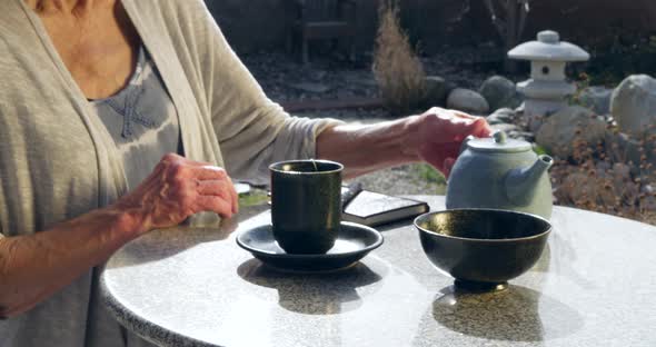 A woman placing an herbal tea bag into a cup and pouring hot water from a teapot then letting it ste