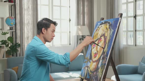 Side View Of An Asian Artist Man Holding Paintbrush And Painting A Girl's Ear On The Canvas