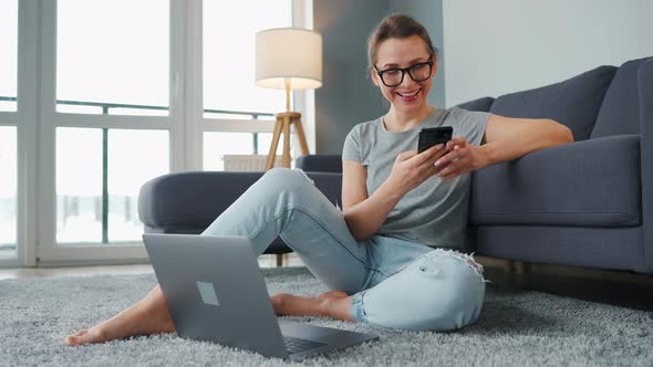 Casually Dressed Woman Sitting on Carpet with Laptop and Smartphone and Working in Cozy Room