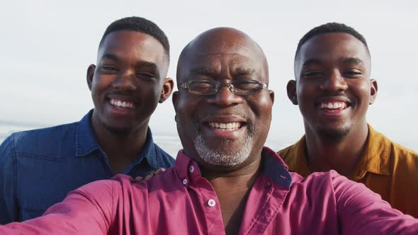 Smiling african american senior father and twin teenage sons standing on a beach taking a selfie