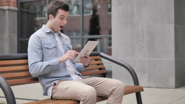 Casual Young Man Sitting Outdoor and Cheering Success on Tablet