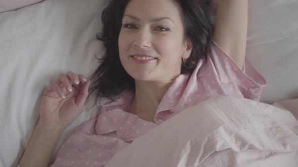 Close-up of Positive Caucasian Brunette Woman Lying in Bed in the Morning. Smiling Happy Lady Waking