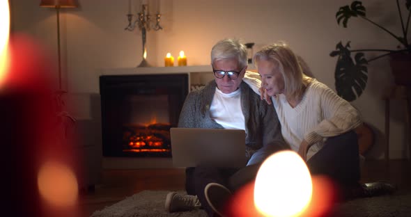 Senior Couple Browsing Internet Together Sitting Near Fireplace on Floor