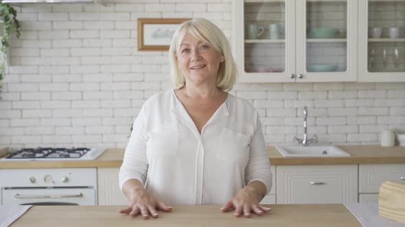 Positive Mature Woman Looking at the Camera Smiling While Standing in the Modern Kitchen. The Senior