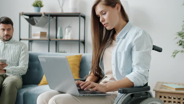 Disabled Female Freelancer Working with Laptop While Handicapped Man Sitting on Sofa Using