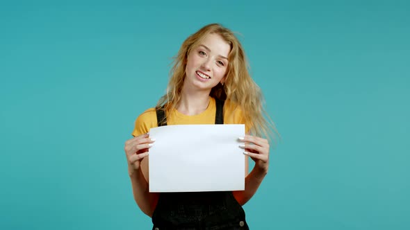 Portrait of Young Woman in Overall Holding White Horizontal A4 Paper Isolated on Blue Studio