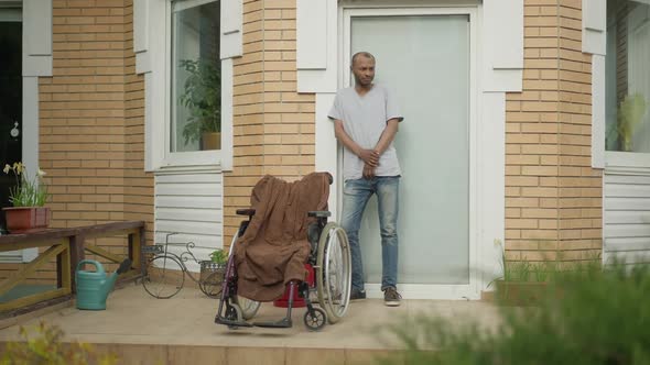 Wide Shot Portrait of Handsome African American Man Standing on Porch with Wheelchair Waiting