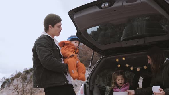 Traveler Family  sitting in the open trunk of a black car with kids near bonfire