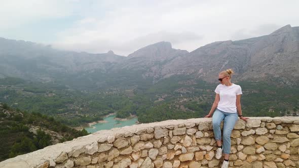 Woman Sitting and Looking at a Beautiful View with a Blue Lake and Mountains From the Wall of an Old