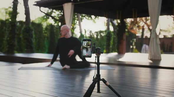 Bald Guy in Black Sport Clothes Performing Yoga Sitting on Mat and Wooden Floor of Inner Yard