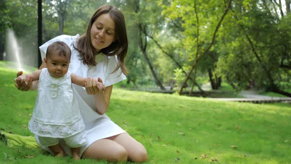 Young Pretty Mother in a White Dress Teaches Her Newborn Daughter To Walk on a Fresh Green Grass in