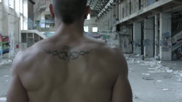 The back of a shirtless personal trainer man does a workout inside an abandoned building.