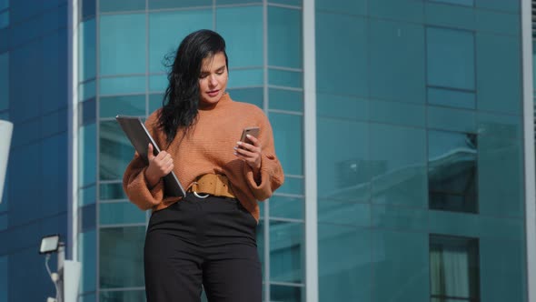 Young Hispanic Businesswoman Stands Backdrop Office Building Looks at Telephone Screen Sending Email