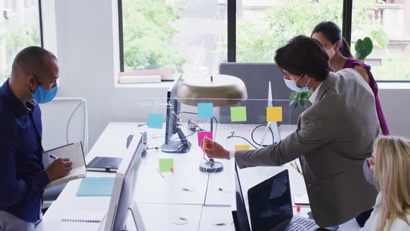 Diverse business colleagues wearing face masks using computers talking in office