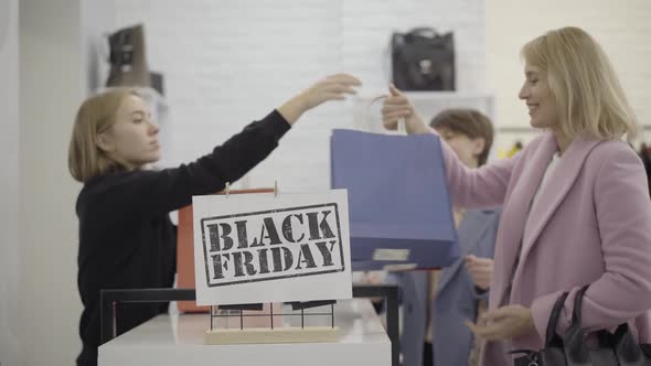 Smiling Shop Assistant Passing Shopping Bags To Happy Clients in Clothing Store. Blurred Caucasian