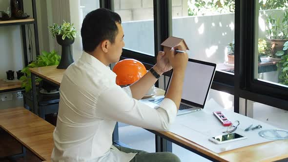 Architect is sitting in front of computer and holding small model of the house.