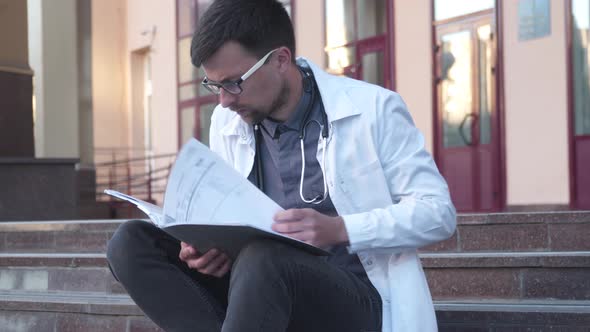 A Medical Student Sits on the Stairs Outside the University and Studies the Medical Report Preparing