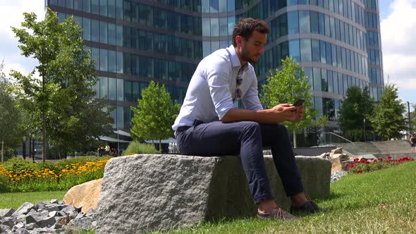 A Young Handsome Businessman Sits in a Park and Works on a Smartphone, an Urban Area