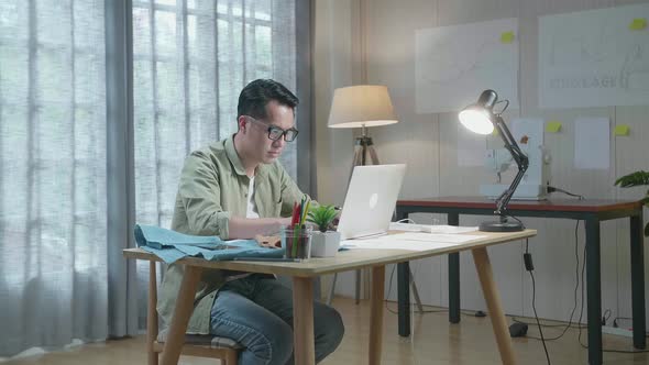 Asian Man Designer With Layout Bond And Fabric Typing On Laptop At The Office