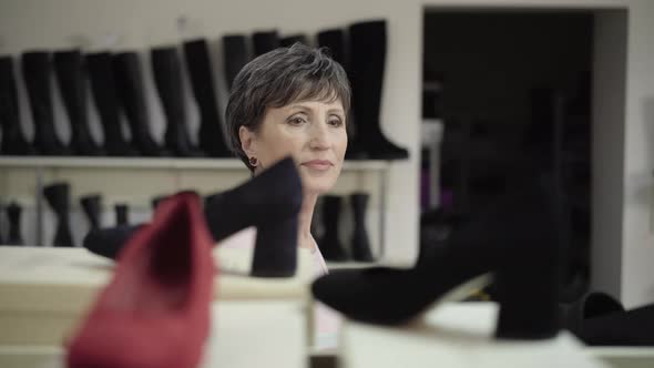 Camera Follows Senior Elegant Woman Walking Along Row of Boots in Shoe Store. Portrait of Confident