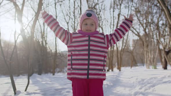 Joyful Little Child Girl Looking at Camera Smiling Dancing on Snowy Road in Winter Park Outdoors